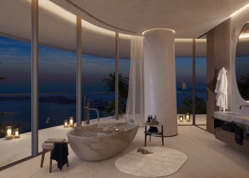 Luxurious bathroom at Como Residences with large floor-to-ceiling windows overlooking a scenic waterfront at dusk. Features an elegant freestanding marble bathtub, a round mirror above a sleek vanity, a bathrobe on a hook, and ambient candle lighting creating a serene atmosphere. | RealEstateMarket