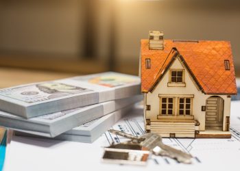 A miniature model of a house with a red roof sits beside two stacks of hundred-dollar bills and a set of house keys, symbolizing the opportunity to buy properties. The items are placed on top of architectural plans, with the background softly blurred to highlight the objects in the foreground. | RealEstateMarket