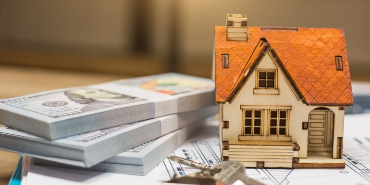 A miniature model of a house with a red roof sits beside two stacks of hundred-dollar bills and a set of house keys, symbolizing the opportunity to buy properties. The items are placed on top of architectural plans, with the background softly blurred to highlight the objects in the foreground. | RealEstateMarket