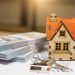 A miniature model of a house with a red roof sits beside two stacks of hundred-dollar bills and a set of house keys, symbolizing the opportunity to buy properties. The items are placed on top of architectural plans, with the background softly blurred to highlight the objects in the foreground. | RealEstateMarket