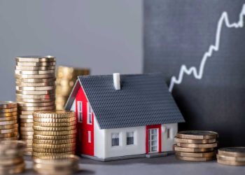 Stacks of gold coins surround a small model house with a red roof, in front of a blackboard featuring an upward-trending graph, serving as the perfect investment guide for real estate and financial growth. | RealEstateMarket