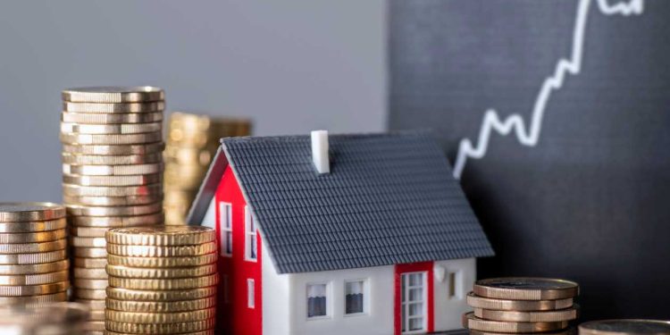 Stacks of gold coins surround a small model house with a red roof, in front of a blackboard featuring an upward-trending graph, serving as the perfect investment guide for real estate and financial growth. | RealEstateMarket