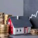 Stacks of gold coins surround a small model house with a red roof, in front of a blackboard featuring an upward-trending graph, serving as the perfect investment guide for real estate and financial growth. | RealEstateMarket