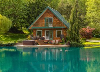 A charming wooden cabin with a green roof sits peacefully beside a vibrant turquoise pond, making it an ideal property investment in North America. The cabin features a balcony, spacious deck, and hot tub. Surrounded by lush green trees and shrubs, the serene setting is enhanced by two ducks swimming in the pond. | RealEstateMarket