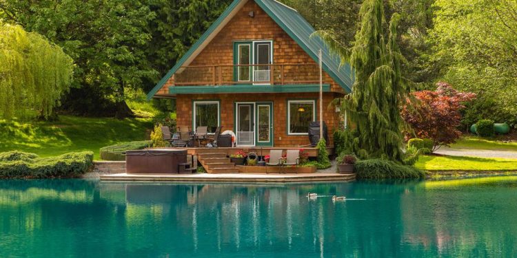 A charming wooden cabin with a green roof sits peacefully beside a vibrant turquoise pond, making it an ideal property investment in North America. The cabin features a balcony, spacious deck, and hot tub. Surrounded by lush green trees and shrubs, the serene setting is enhanced by two ducks swimming in the pond. | RealEstateMarket