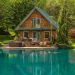 A charming wooden cabin with a green roof sits peacefully beside a vibrant turquoise pond, making it an ideal property investment in North America. The cabin features a balcony, spacious deck, and hot tub. Surrounded by lush green trees and shrubs, the serene setting is enhanced by two ducks swimming in the pond. | RealEstateMarket