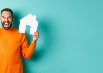 A man in an orange sweater smiles, pondering how much to afford a 500K house, as he holds a paper house in one hand and cash in the other, standing against a teal background. | RealEstateMarket