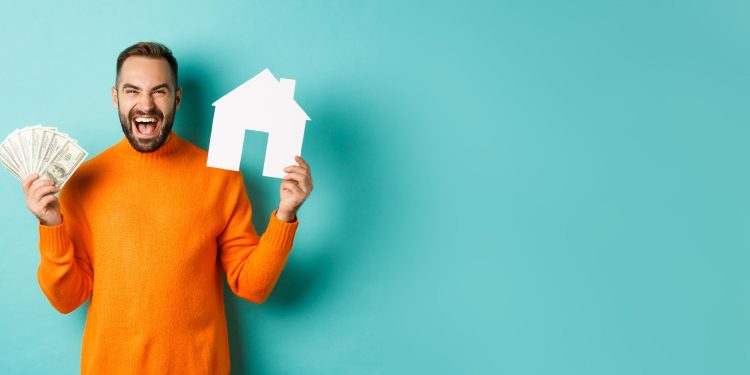 A man in an orange sweater smiles, pondering how much to afford a 500K house, as he holds a paper house in one hand and cash in the other, standing against a teal background. | RealEstateMarket