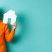 A man in an orange sweater smiles, pondering how much to afford a 500K house, as he holds a paper house in one hand and cash in the other, standing against a teal background. | RealEstateMarket