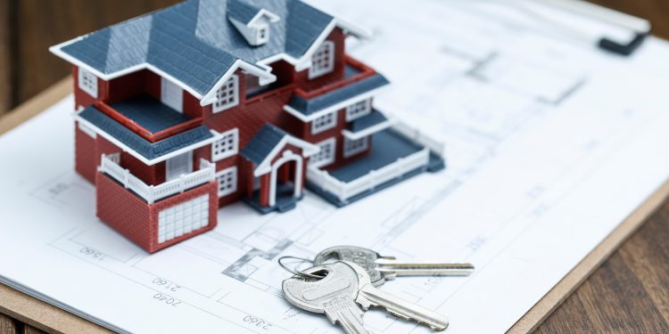 A small model of a red house with a gray roof sits on a clipboard holding blueprints, symbolizing the concept of fractional real estate. Two metallic keys rest on the plans, atop a wooden table that adds warmth to this contemporary investment scene. | RealEstateMarket