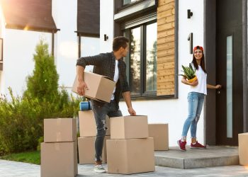 A man and a woman move boxes into a modern house on a sunny day. With "The Quick and Easy Guide to Moving House Without Stress" in mind, the man carries boxes while the woman, in a red cap, holds a plant and opens the door. Luggage is visible nearby. | RealEstateMarket