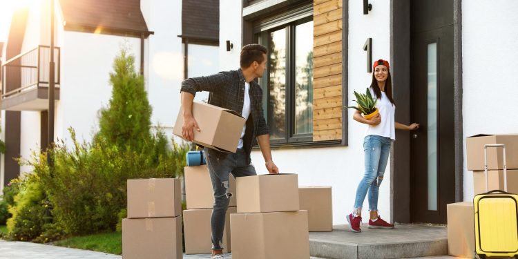 A man and a woman move boxes into a modern house on a sunny day. With "The Quick and Easy Guide to Moving House Without Stress" in mind, the man carries boxes while the woman, in a red cap, holds a plant and opens the door. Luggage is visible nearby. | RealEstateMarket