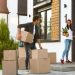 A man and a woman move boxes into a modern house on a sunny day. With "The Quick and Easy Guide to Moving House Without Stress" in mind, the man carries boxes while the woman, in a red cap, holds a plant and opens the door. Luggage is visible nearby. | RealEstateMarket
