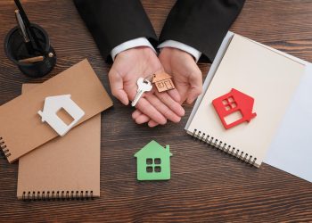 A person in a suit holds a house-shaped keychain and a key above a wooden desk with notepads, a pen holder, and house-shaped cutouts, highlighting the decision between renting vs flipping properties. | RealEstateMarket