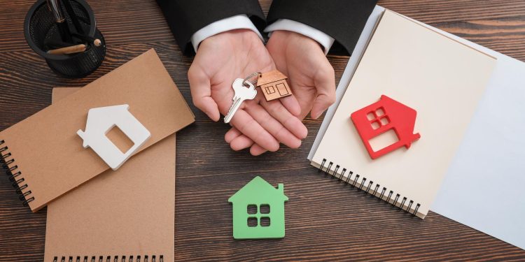 A person in a suit holds a house-shaped keychain and a key above a wooden desk with notepads, a pen holder, and house-shaped cutouts, highlighting the decision between renting vs flipping properties. | RealEstateMarket