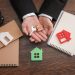 A person in a suit holds a house-shaped keychain and a key above a wooden desk with notepads, a pen holder, and house-shaped cutouts, highlighting the decision between renting vs flipping properties. | RealEstateMarket