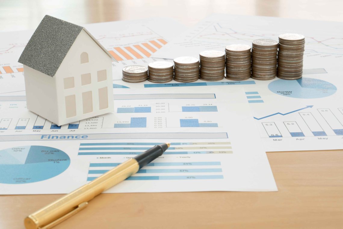 A small model house, stacks of coins, and a pen rest on financial charts and graphs, symbolizing building a profitable real estate portfolio through investment, financial growth, and analysis. | RealEstateMarket