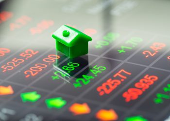 A green toy house sits on a digital financial screen displaying red and green stock numbers and arrows, symbolizing Real Estate ROI. | RealEstateMarket