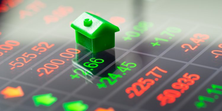 A green toy house sits on a digital financial screen displaying red and green stock numbers and arrows, symbolizing Real Estate ROI. | RealEstateMarket