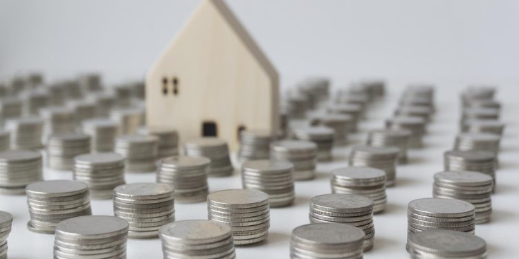 A small wooden house model is surrounded by numerous neatly stacked piles of silver coins on a white surface, suggesting themes of savings, finance, or investment in property. | RealEstateMarket