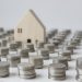 A small wooden house model is surrounded by numerous neatly stacked piles of silver coins on a white surface, suggesting themes of savings, finance, or investment in property. | RealEstateMarket