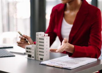 A person in a red blazer sits at a desk with small building models, a contract, and a pen, gesturing with one hand while holding a pen in the other. | RealEstateMarket