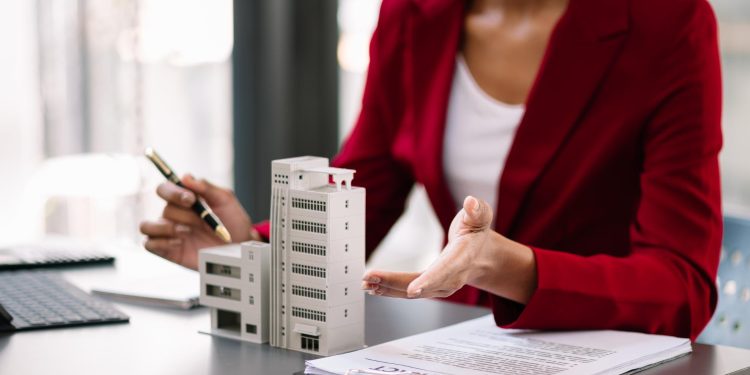 A person in a red blazer sits at a desk with small building models, a contract, and a pen, gesturing with one hand while holding a pen in the other. | RealEstateMarket