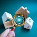 A hand holds a magnifying glass over small model houses, focusing on digital real estate and financial icons, representing property search, investment, and market analysis. | RealEstateMarket