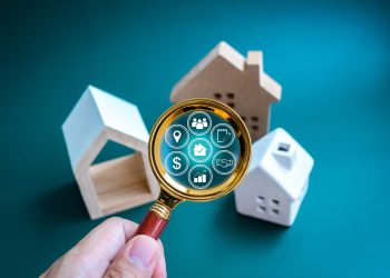 A hand holds a magnifying glass over small model houses, revealing digital icons related to real estate, such as a house, dollar sign, location pin, and charts, on a teal background. | RealEstateMarket