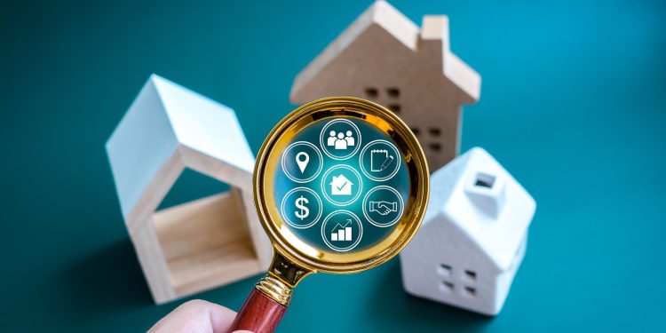 A hand holds a magnifying glass over small model houses, revealing digital icons related to real estate, such as a house, dollar sign, location pin, and charts, on a teal background. | RealEstateMarket
