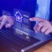 A person uses a laptop, interacting with floating digital icons of house listings, suggesting online real estate or property search. The scene has a blue tint and shows a hand touching a virtual screen. | RealEstateMarket