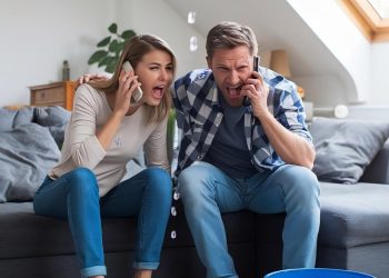 A worried couple sits on a couch, talking on their phones while water leaks from the ceiling into a blue bucket on the floor, causing a large puddle in their living room. | RealEstateMarket