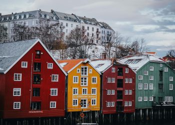 Colorful wooden buildings in red, yellow, and green stand by a waterfront, with a larger white apartment building and bare trees on a hill in the background under a cloudy sky. | RealEstateMarket