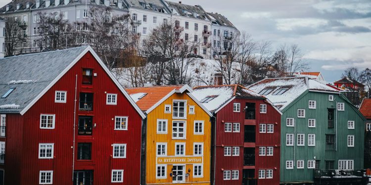 Colorful wooden buildings in red, yellow, and green stand by a waterfront, with a larger white apartment building and bare trees on a hill in the background under a cloudy sky. | RealEstateMarket