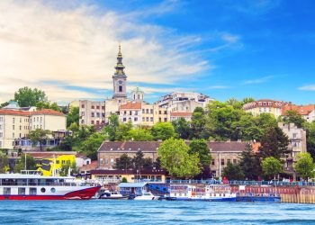 A riverside view of Belgrade, Serbia, featuring boats docked along the water, historic buildings, greenery, and the prominent bell tower of the Church of St. Michael in the background under a blue sky. | RealEstateMarket