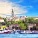 A riverside view of Belgrade, Serbia, featuring boats docked along the water, historic buildings, greenery, and the prominent bell tower of the Church of St. Michael in the background under a blue sky. | RealEstateMarket