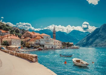 A coastal village with red-roofed buildings lines a turquoise bay, surrounded by mountains. Small boats float on the water under a bright, clear sky with scattered clouds. A stone path follows the waterfront. | RealEstateMarket