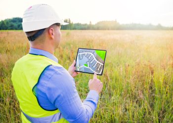 A person wearing a safety helmet and reflective vest stands in a field, holding a tablet displaying a digital map with a marked location. Trees and tall grass are visible in the background under a bright sky. | RealEstateMarket
