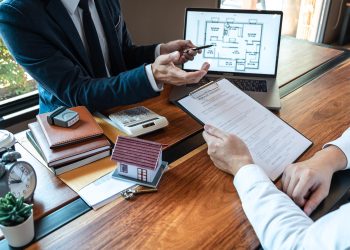Two people in business attire sit at a desk discussing documents. A small model house, calculator, notepad, and laptop showing a floor plan are on the table, suggesting a real estate or property discussion. | RealEstateMarket