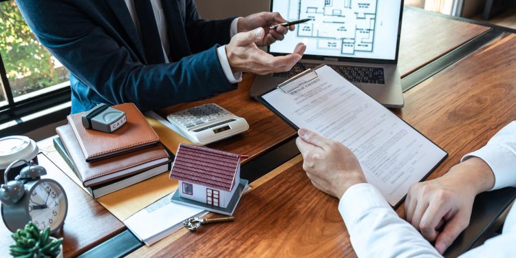 Two people in business attire sit at a desk discussing documents. A small model house, calculator, notepad, and laptop showing a floor plan are on the table, suggesting a real estate or property discussion. | RealEstateMarket