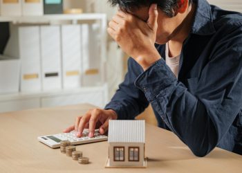 A person sits at a desk with their head in their hand, looking stressed while using a calculator. There are coins stacked on the table and a small model house, suggesting financial concerns or housing expenses. | RealEstateMarket