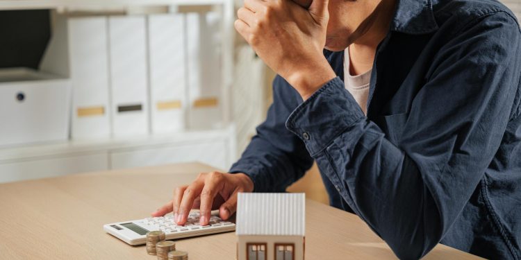 A person sits at a desk with their head in their hand, looking stressed while using a calculator. There are coins stacked on the table and a small model house, suggesting financial concerns or housing expenses. | RealEstateMarket