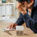 A person sits at a desk with their head in their hand, looking stressed while using a calculator. There are coins stacked on the table and a small model house, suggesting financial concerns or housing expenses. | RealEstateMarket