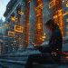 A person sits on stone steps outside a grand, columned building at dusk, using a tablet. Orange digital icons and data graphics float around them, blending technology with the classical architecture. | RealEstateMarket