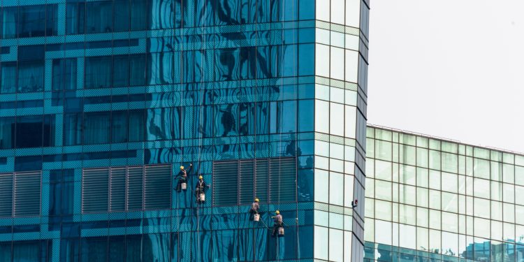 Four workers, equipped with safety gear, clean the blue glass windows of a tall modern building, suspended by ropes. The building reflects distorted images of nearby structures. | RealEstateMarket