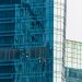 Four workers, equipped with safety gear, clean the blue glass windows of a tall modern building, suspended by ropes. The building reflects distorted images of nearby structures. | RealEstateMarket