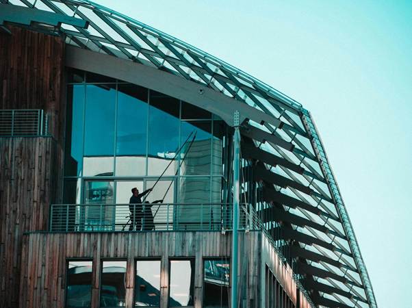A person stands on a balcony, cleaning large glass windows of a modern building with a long pole, under a clear blue sky. The building features wooden and glass architectural elements. | RealEstateMarket