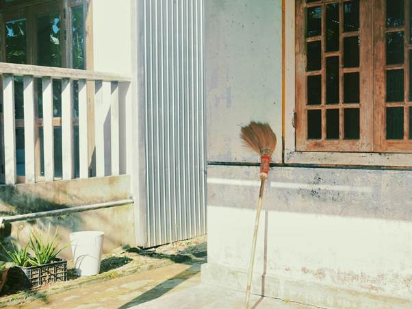A broom leans against a sunlit wall next to a wooden window. Nearby, there’s a white railing, green plants, and a white bucket. The overall scene appears calm and bright with natural light. | RealEstateMarket