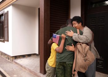 A family embraces outside a house; the child holds a small American flag, welcoming home a loved one in military uniform. | RealEstateMarket