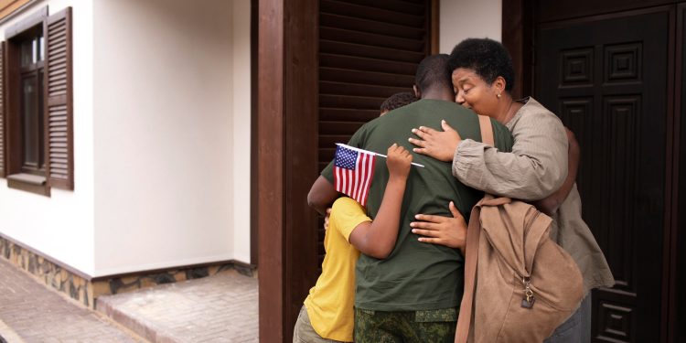 A family embraces outside a house; the child holds a small American flag, welcoming home a loved one in military uniform. | RealEstateMarket
